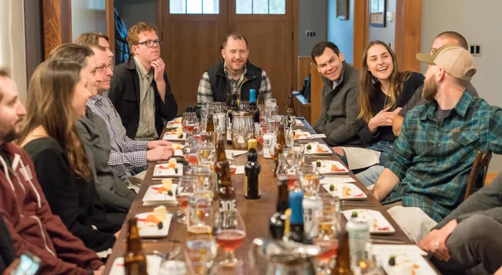 A group of adults talking and laughing around a long dinner table with drinks and small plates