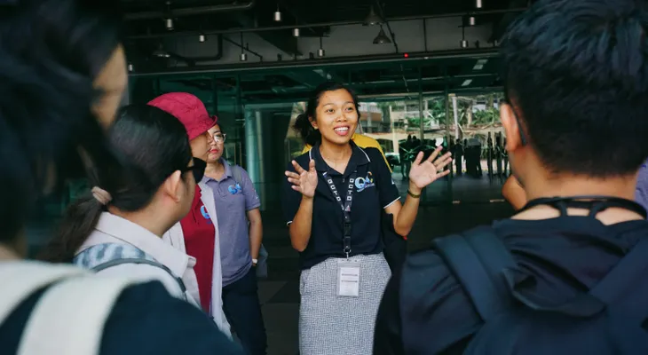 A smiling tour guide with a lanyard talking to a small group of travelers outside a glass-fronted building