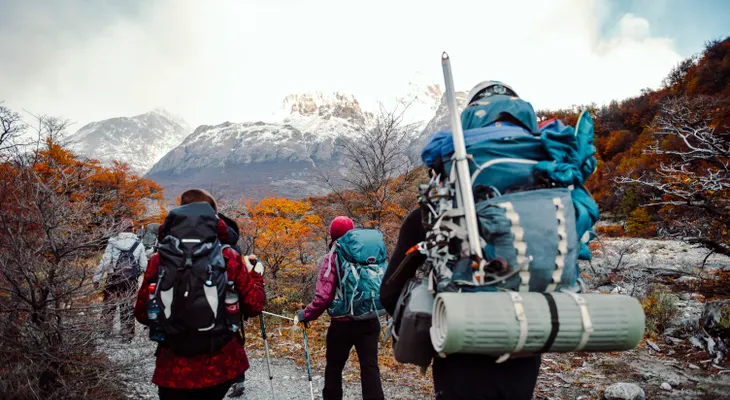 A group of hikers with large backpacks walking a trail through autumn trees toward snowy mountains