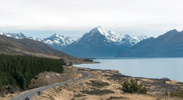 A road running beside a pale blue lake toward snow-capped mountains