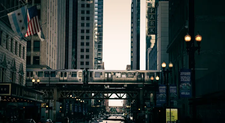A Chicago 'L' train crossing an elevated bridge over a downtown street lined with tall buildings and lamp posts