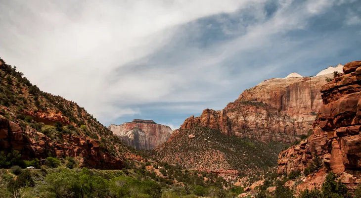 Red and cream sandstone cliffs rising above a green valley in Zion National Park under a streaky sky