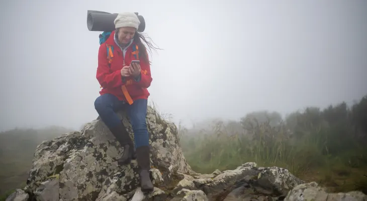 A hiker in a red jacket sitting on a rock in thick fog, checking her phone