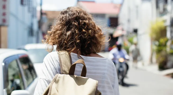 A woman with a canvas backpack walking along a sunny street as a scooter comes toward her