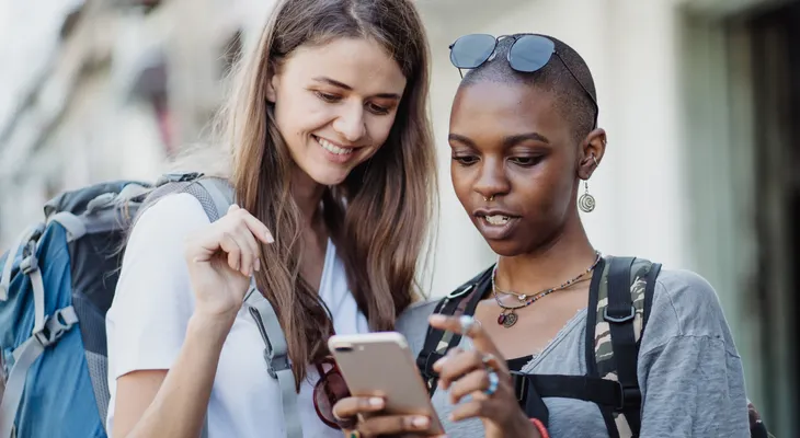 Two backpackers standing close together and smiling at a phone one of them is holding
