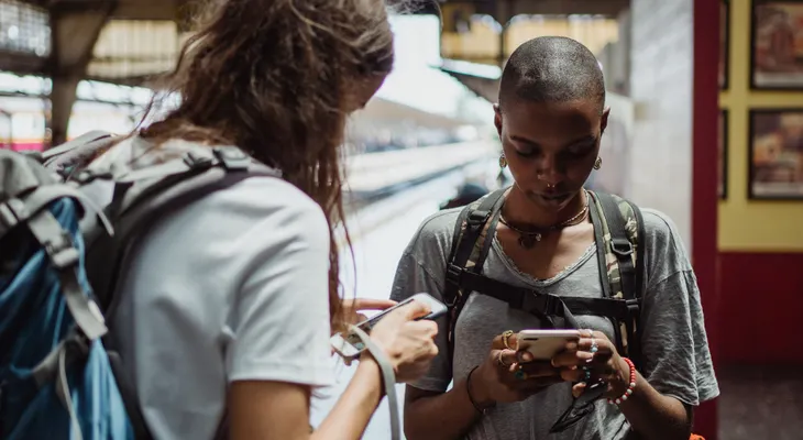 Two travelers with backpacks looking at their phones on a train station platform